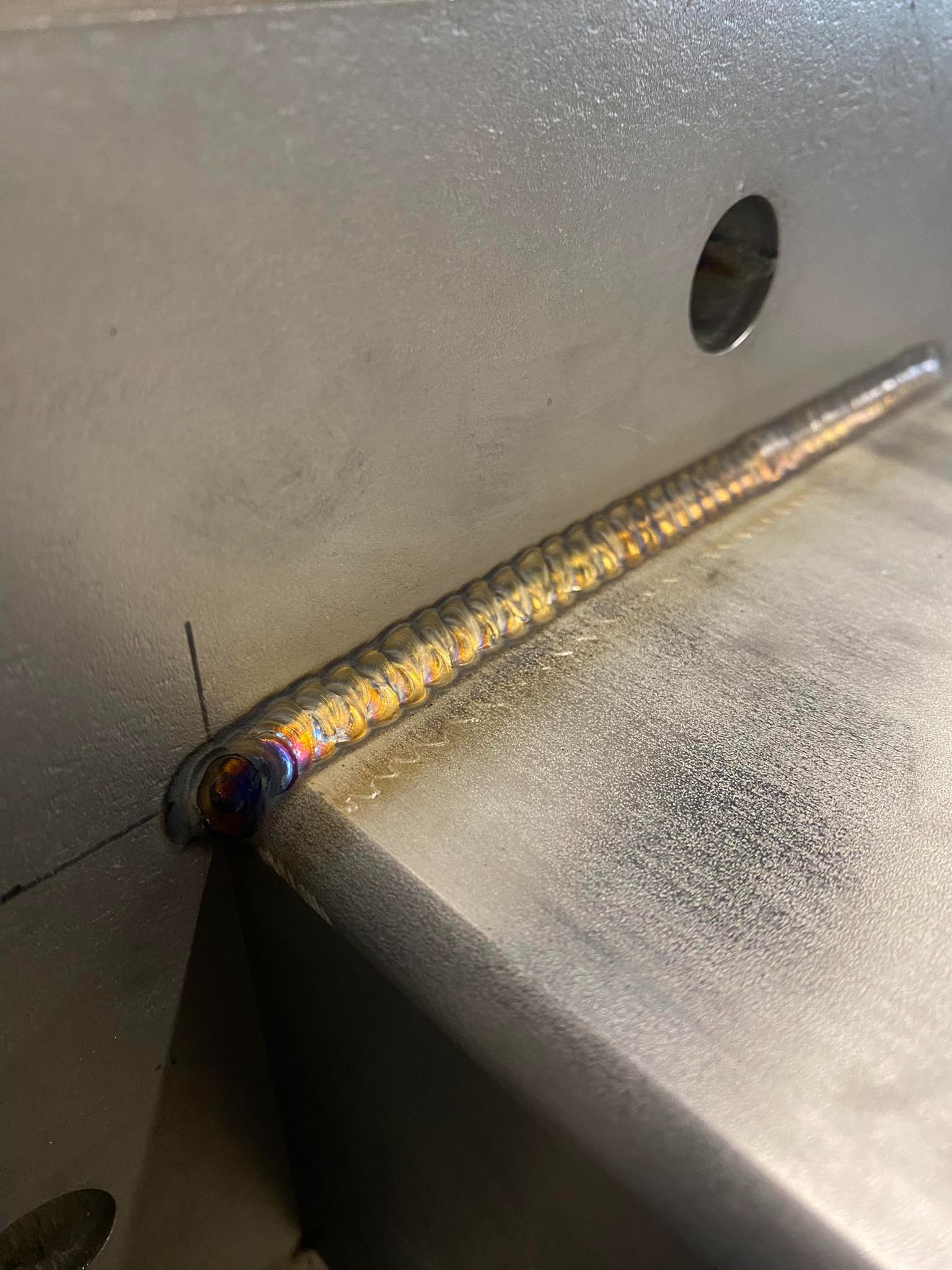 Precyzyjny spaw na metalowej konstrukcji, widoczne tęczowe przebarwienia świadczące o wysokiej jakości wykonania. Detal spoiny z bliska, z otworem w tle.