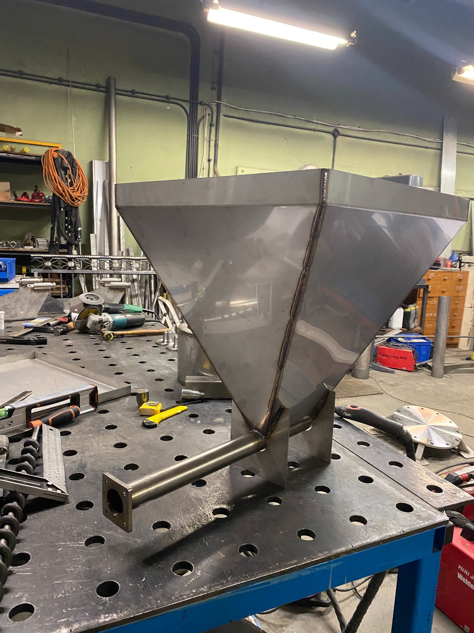 Metalowy zbiornik z rurą w warsztacie, widoczne spawy, narzędzia na perforowanym stole spawalniczym. Element konstrukcyjny w trakcie realizacji.