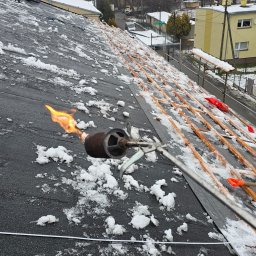 Pro Dach - Praca dekarska w zimowych warunkach: palnik dekarski w akcji na pokrytym śniegiem dachu z papy, w tle zabudowa miejska i drzewa bez liści.