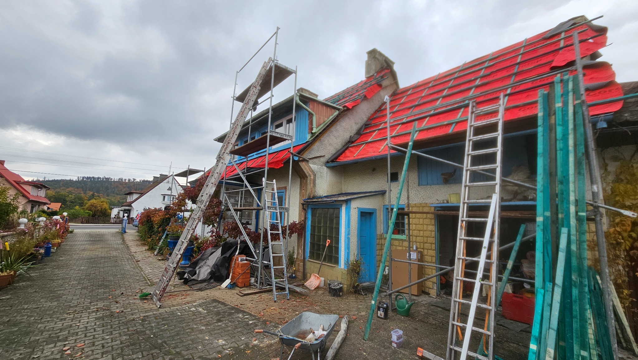 Dom w trakcie wymiany dachu, widoczne rusztowania i czerwona membrana dachowa. Prace dekarskie w toku, zieleń w tle, pochmurne niebo.