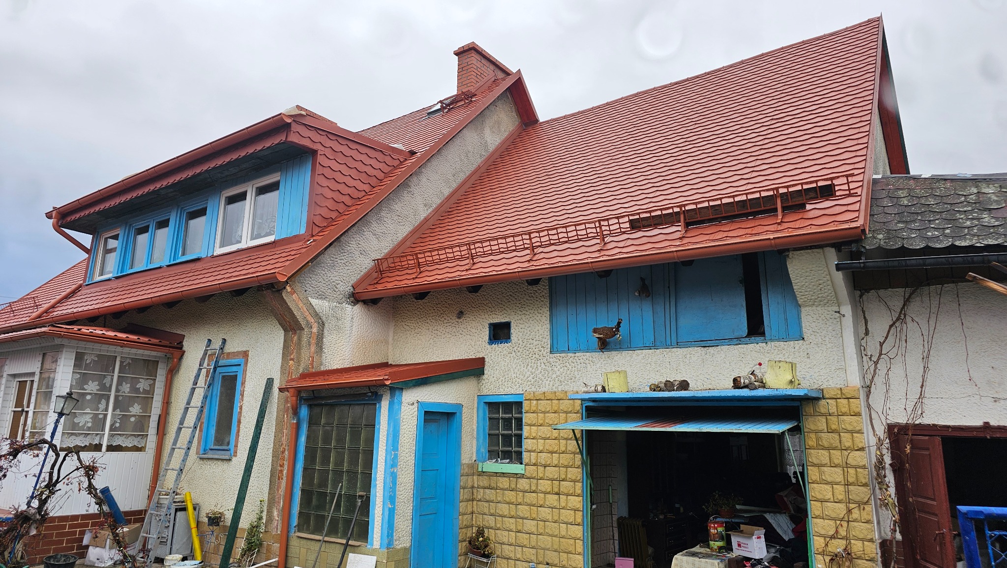 Dom po renowacji dachu z czerwoną dachówką i niebieskimi elementami stolarki okiennej. Widoczne rynny i drabina oparta o ścianę. Ujęcie z dołu, pochmurne niebo.