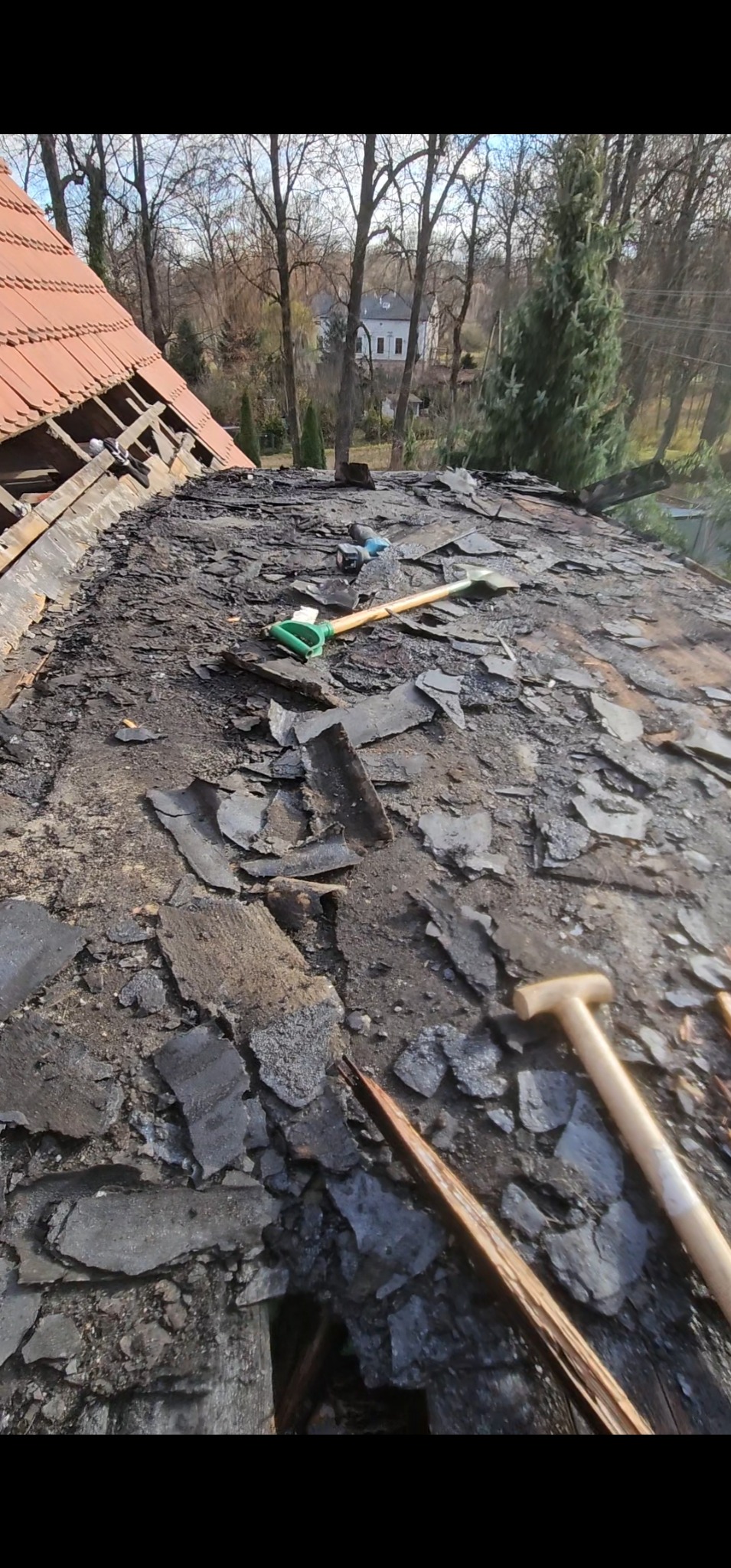 Demontaż starego dachu z papy. Widoczne narzędzia: łopata i młotek dekarski. W tle drzewa i budynek. Fragment dachu z czerwoną dachówką w lewym górnym rogu.
