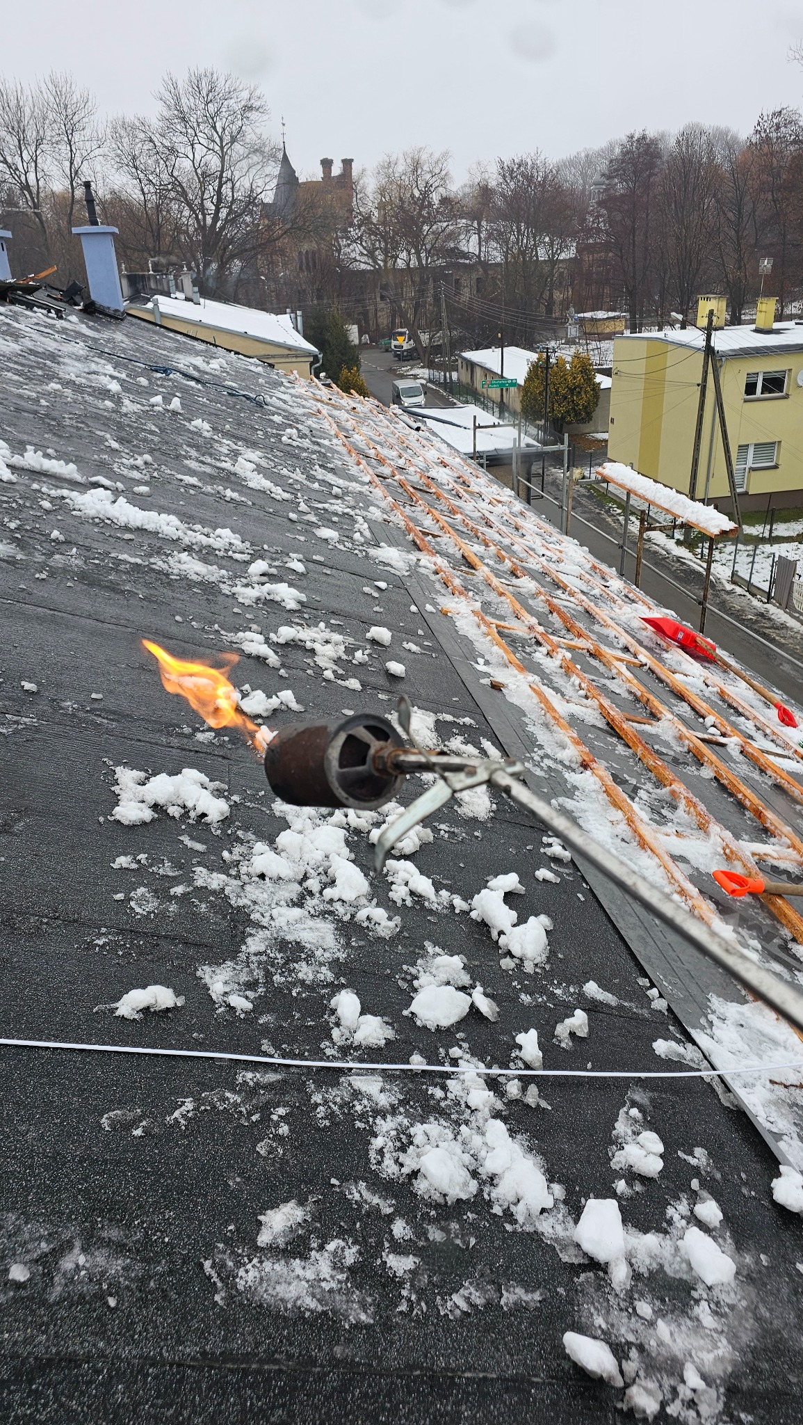 Praca dekarska w zimowych warunkach: palnik dekarski w akcji na pokrytym śniegiem dachu z papy, w tle zabudowa miejska i drzewa bez liści.