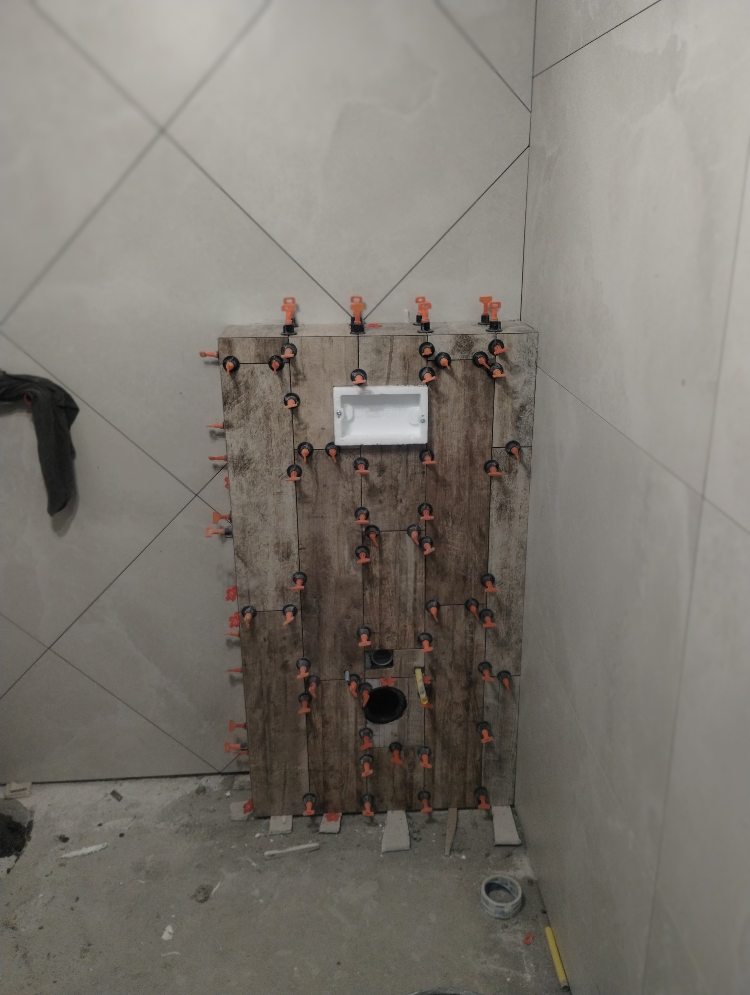 Montaż płytek imitujących drewno na stelażu WC w łazience. Widoczne klipsy poziomujące i otwór na odpływ. Ściany wyłożone jasnymi płytkami z delikatnym wzorem.