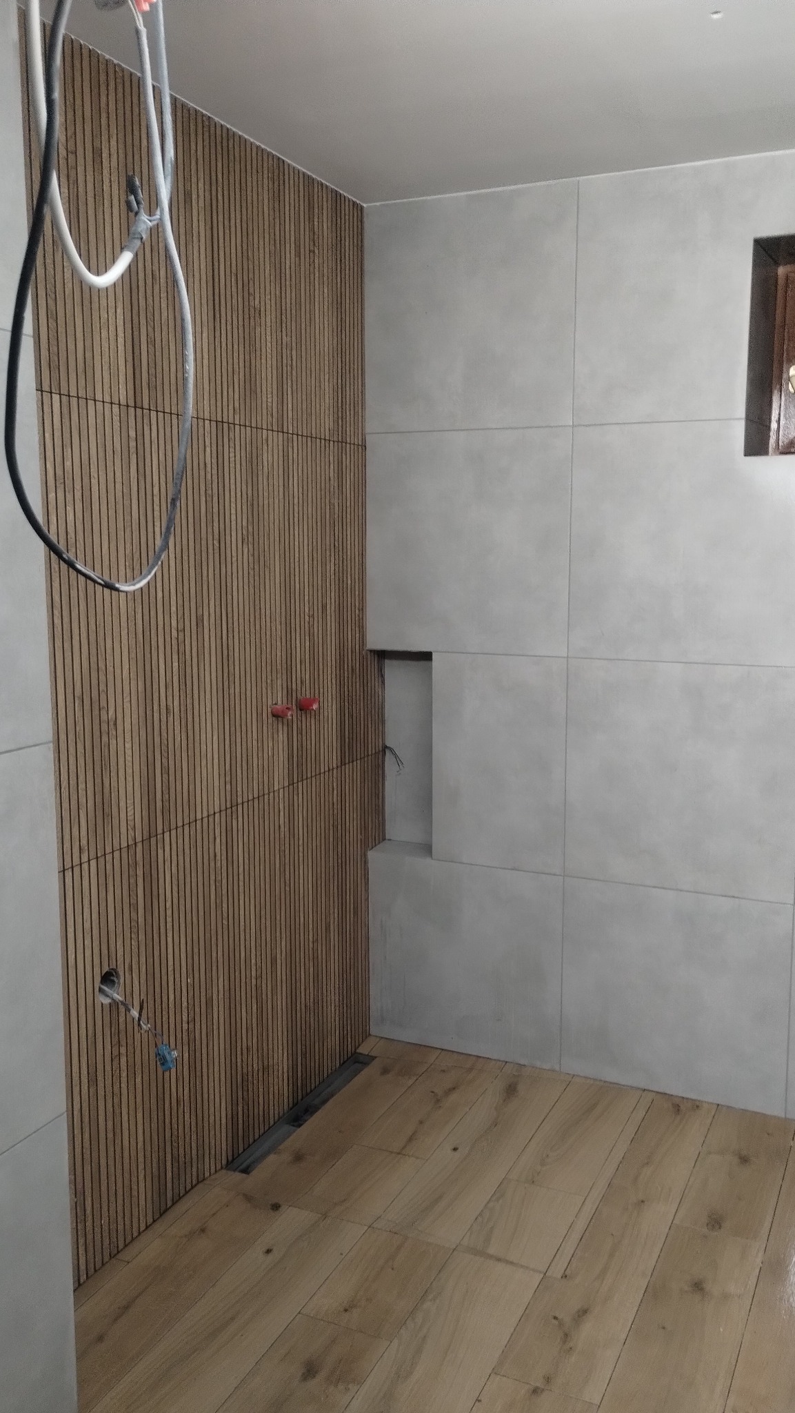 Wykończona łazienka z drewnianą ścianą z lameli, szarymi płytkami i drewnopodobną podłogą. Widoczne instalacje i odpływ liniowy.