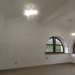 budujdomek - Białe, małe płytki na ścianie w jasnym pomieszczeniu z dwoma łukowatymi oknami z drewnianą ramą. Widoczne przygotowane punkty instalacji elektrycznej.