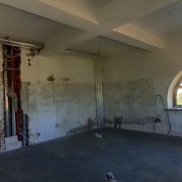 budujdomek - Wnętrze w trakcie remontu: odsłonięte instalacje, surowe ściany z widocznymi śladami prac, okno łukowe z widokiem na zieleń. Widoczne rury i kable.