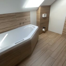 budujdomek - Jasna łazienka na poddaszu z wanną narożną i drewnopodobnymi płytkami na ścianach i podłodze. Widoczne otwory na armaturę i wentylacja.