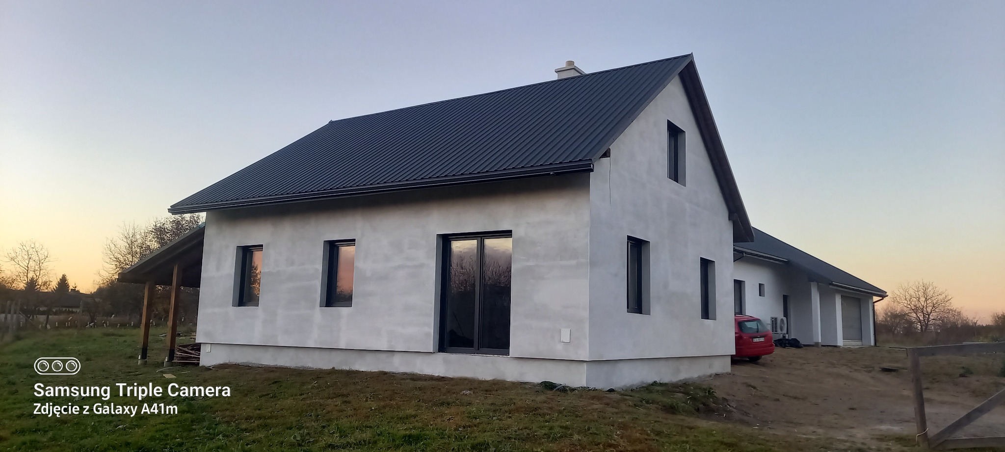 Nowoczesny dom jednorodzinny z ciemnym dachem i jasną elewacją w trakcie wykończenia, widoczny fragment czerwonego samochodu i otoczenie działki.