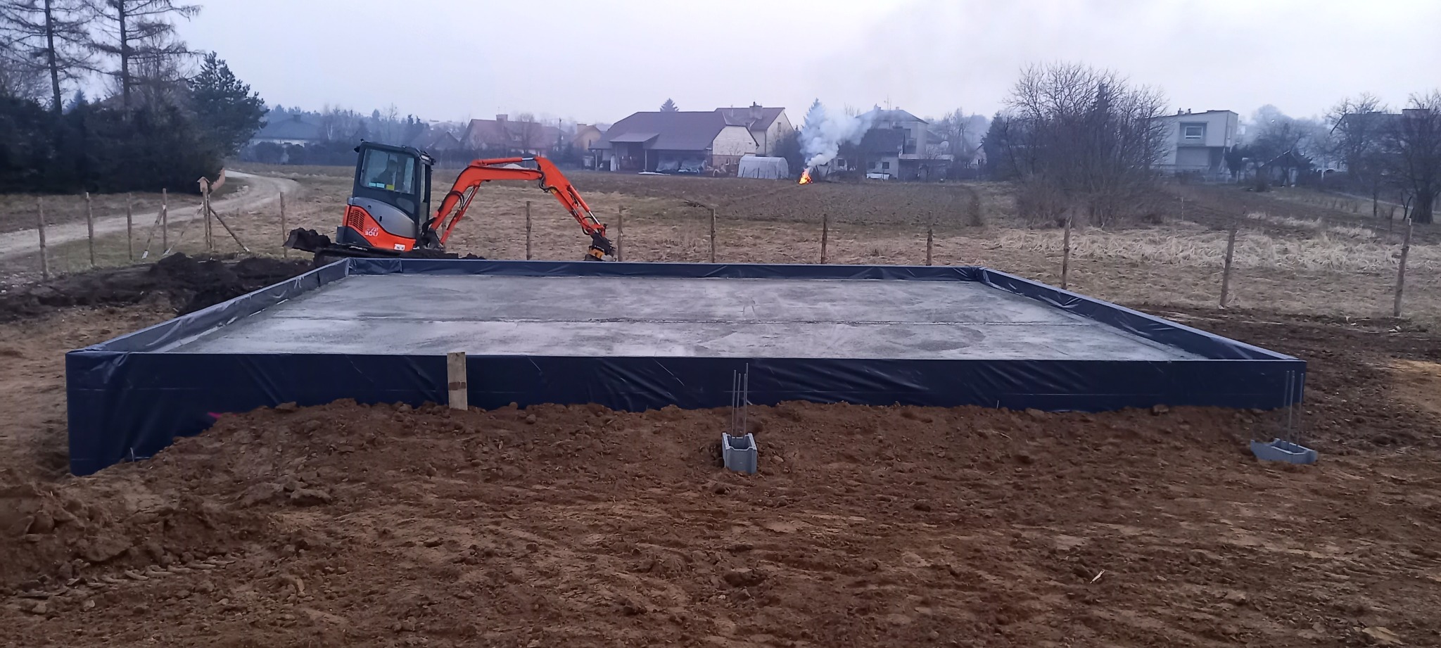 Wylana płyta fundamentowa zabezpieczona folią, z widocznym mini-koparką w tle. Na drugim planie zabudowa wiejska i dym.