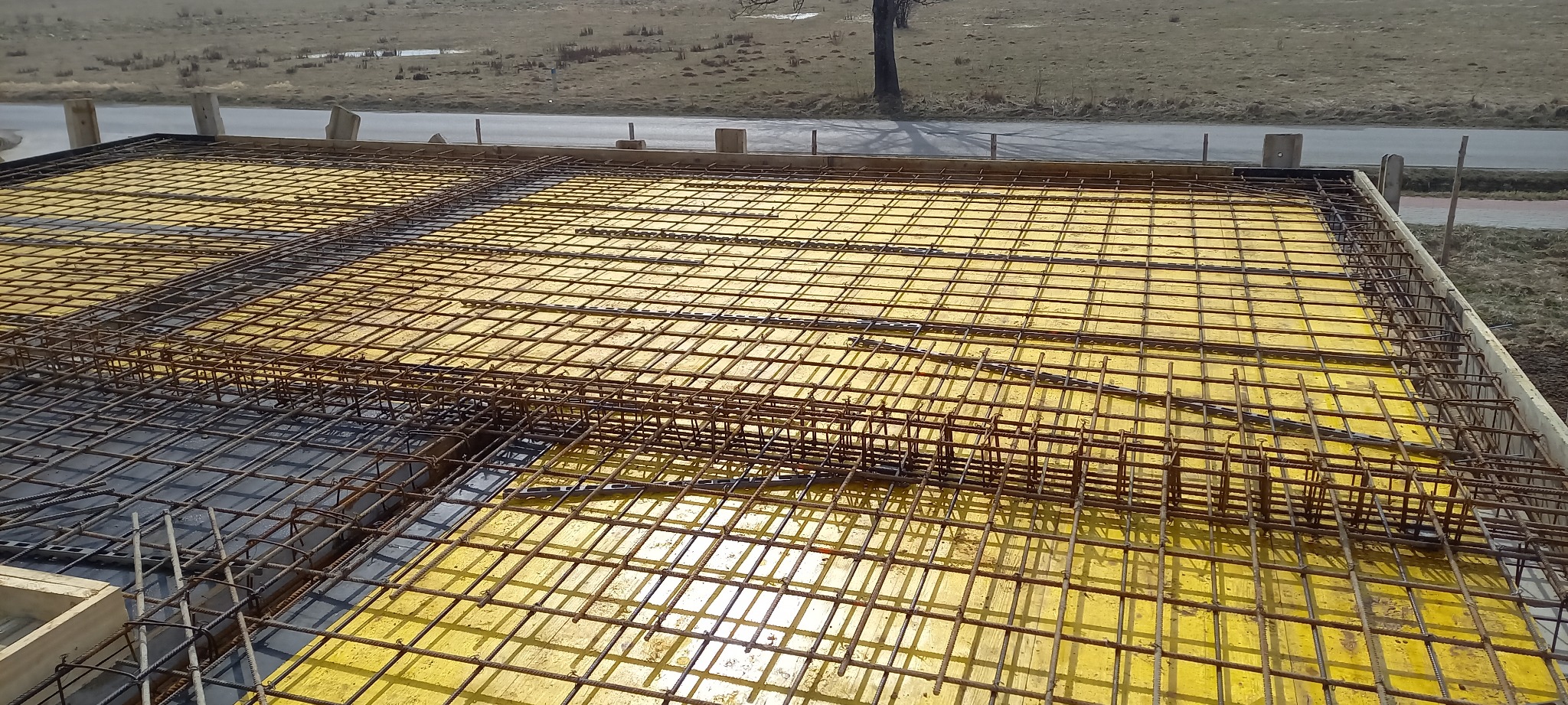 Zbrojenie stropu na żółtym szalunku, widok z góry. Metalowa siatka zbrojeniowa ułożona na deskach szalunkowych. Prace budowlane w toku, przygotowanie do zalania betonem.