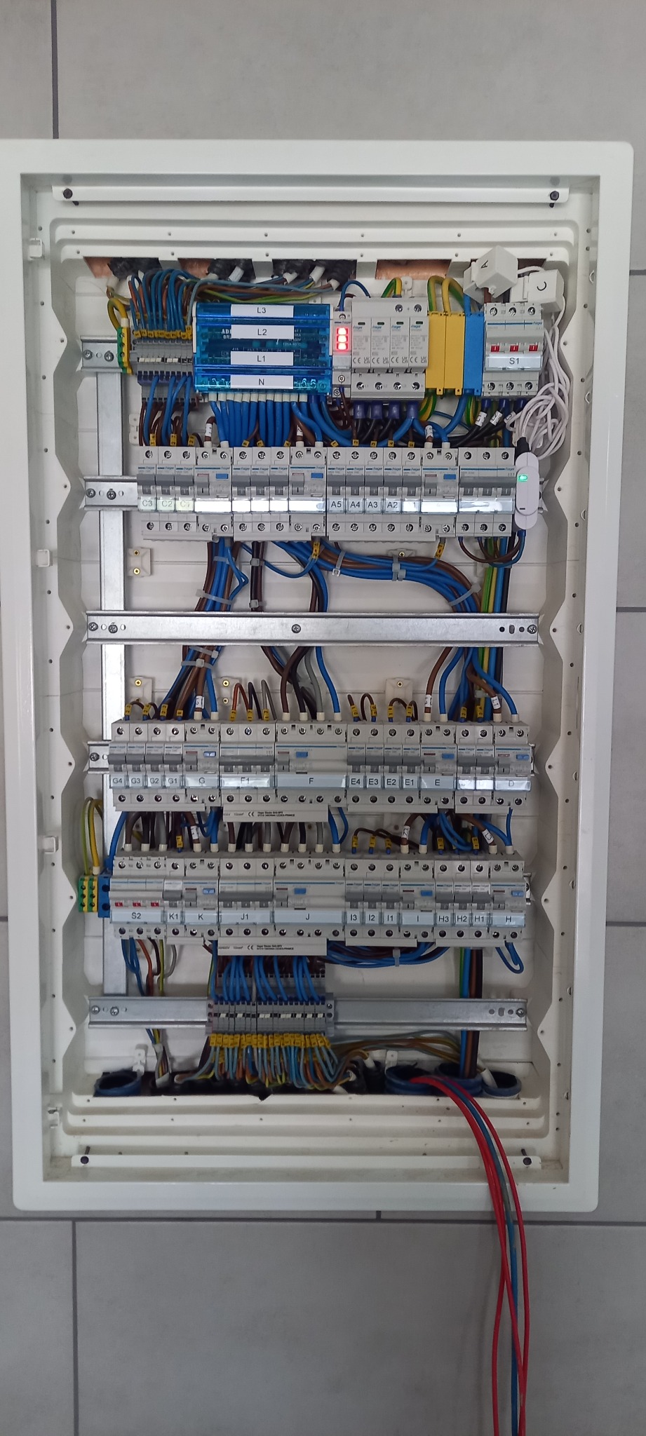 Rozdzielnica elektryczna w białej obudowie, z widocznymi bezpiecznikami, przewodami w kolorach niebieskim, brązowym i żółto-zielonym. Instalacja po montażu.