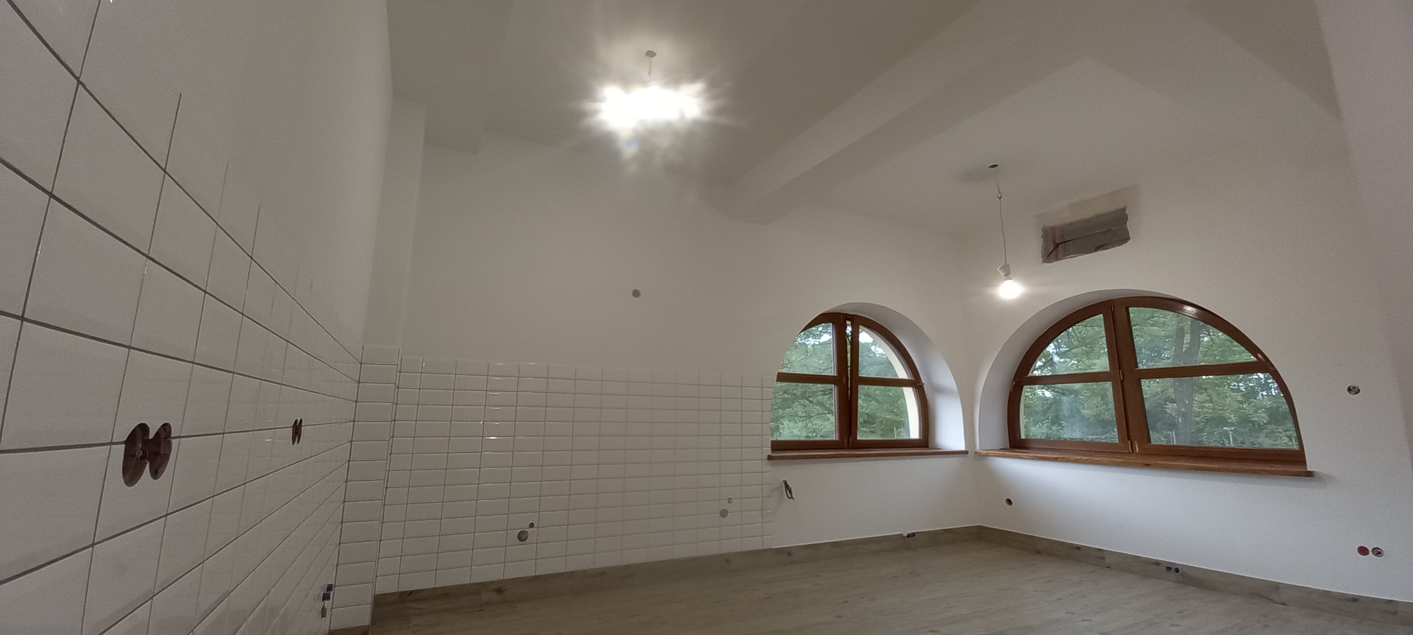 Białe, małe płytki na ścianie w jasnym pomieszczeniu z dwoma łukowatymi oknami z drewnianą ramą. Widoczne przygotowane punkty instalacji elektrycznej.