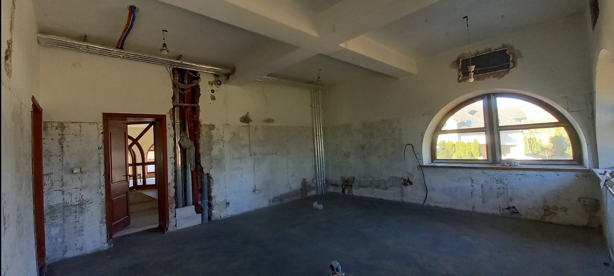 Wnętrze w trakcie remontu: odsłonięte instalacje, surowe ściany z widocznymi śladami prac, okno łukowe z widokiem na zieleń. Widoczne rury i kable.