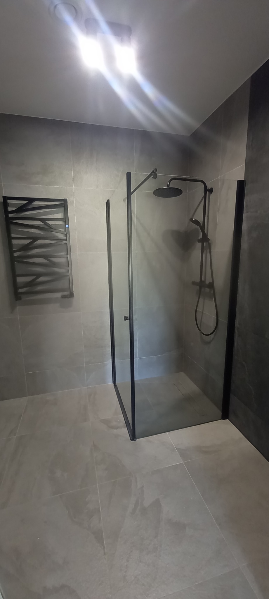 Nowoczesna łazienka z szarymi płytkami, kabiną prysznicową typu walk-in z czarnymi profilami i grzejnikiem drabinkowym o geometrycznym wzorze.