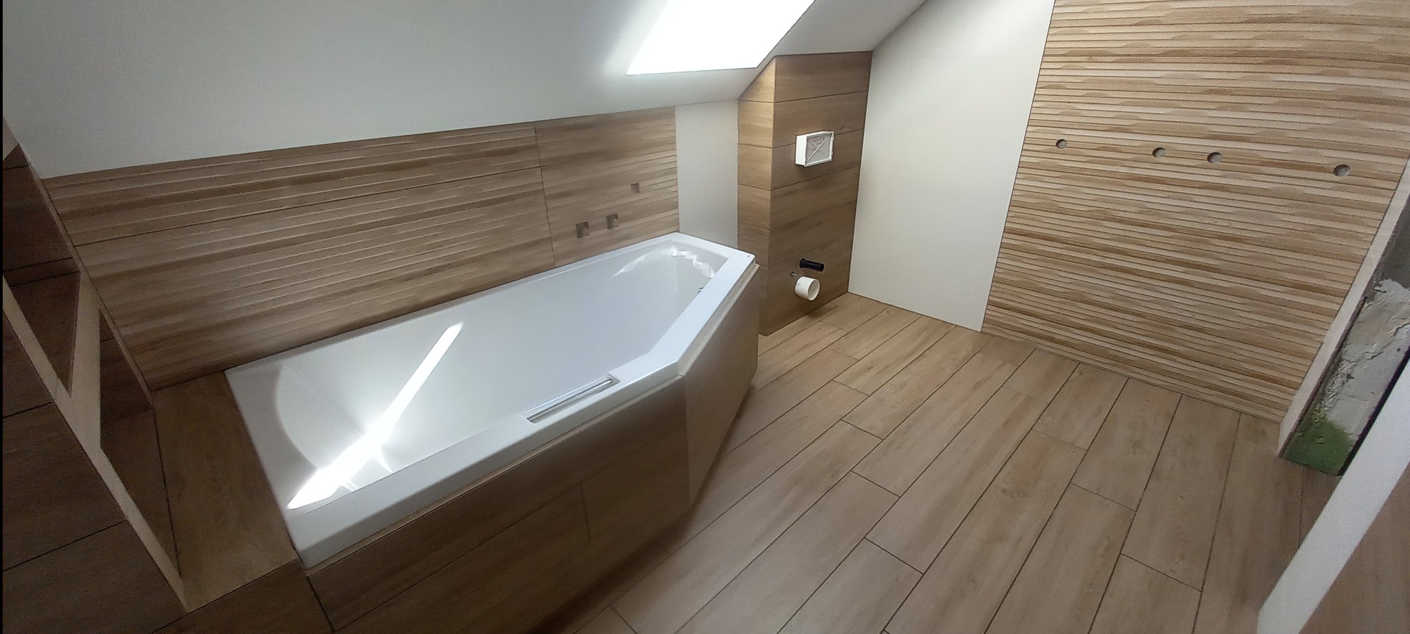 Jasna łazienka na poddaszu z wanną narożną i drewnopodobnymi płytkami na ścianach i podłodze. Widoczne otwory na armaturę i wentylacja.