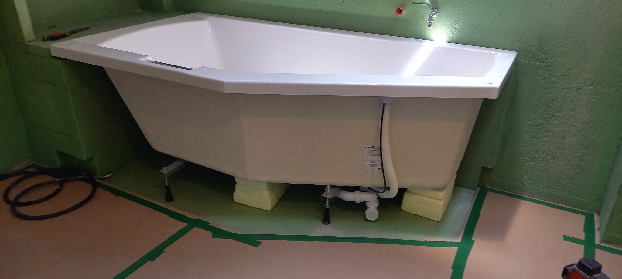 Biała wanna z hydromasażem, ustawiona na żółtych blokach w trakcie montażu w zielonej łazience. Widoczne rury i elementy instalacji wodnej.