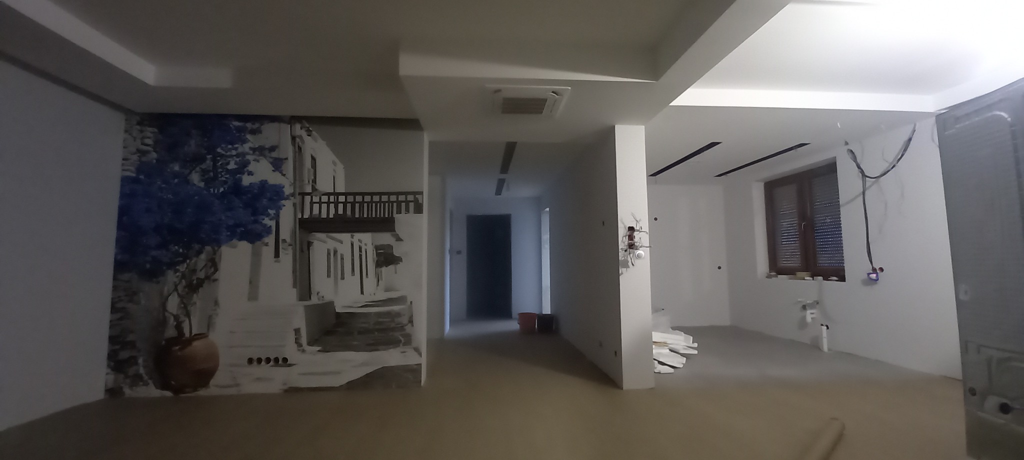 Wnętrze w trakcie remontu: ściana z fototapetą przedstawiającą grecką architekturę, surowy stan wykończenia, widoczne instalacje elektryczne i materiały budowlane.