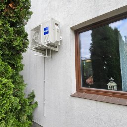 Hydroklima Adrian Pempczak - Biała jednostka zewnętrzna klimatyzacji Haier zamontowana na elewacji domu obok okna, z widoczną instalacją i zielonym krzewem z boku.