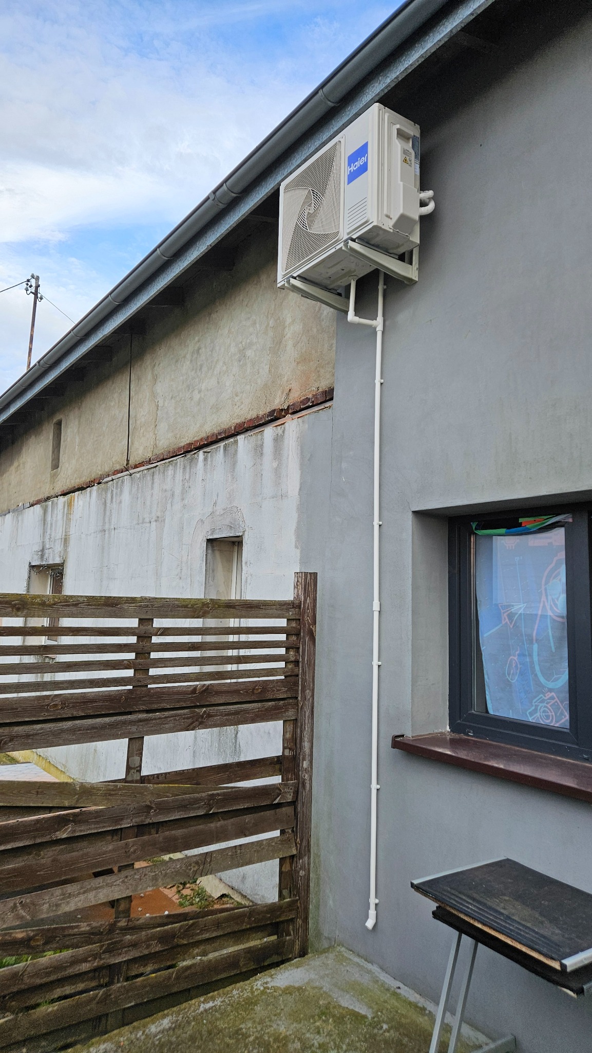 Biały klimatyzator Haier zamontowany na szarej ścianie budynku, widoczne rury odprowadzające skropliny. W tle drewniany płot i fragment drugiego budynku.