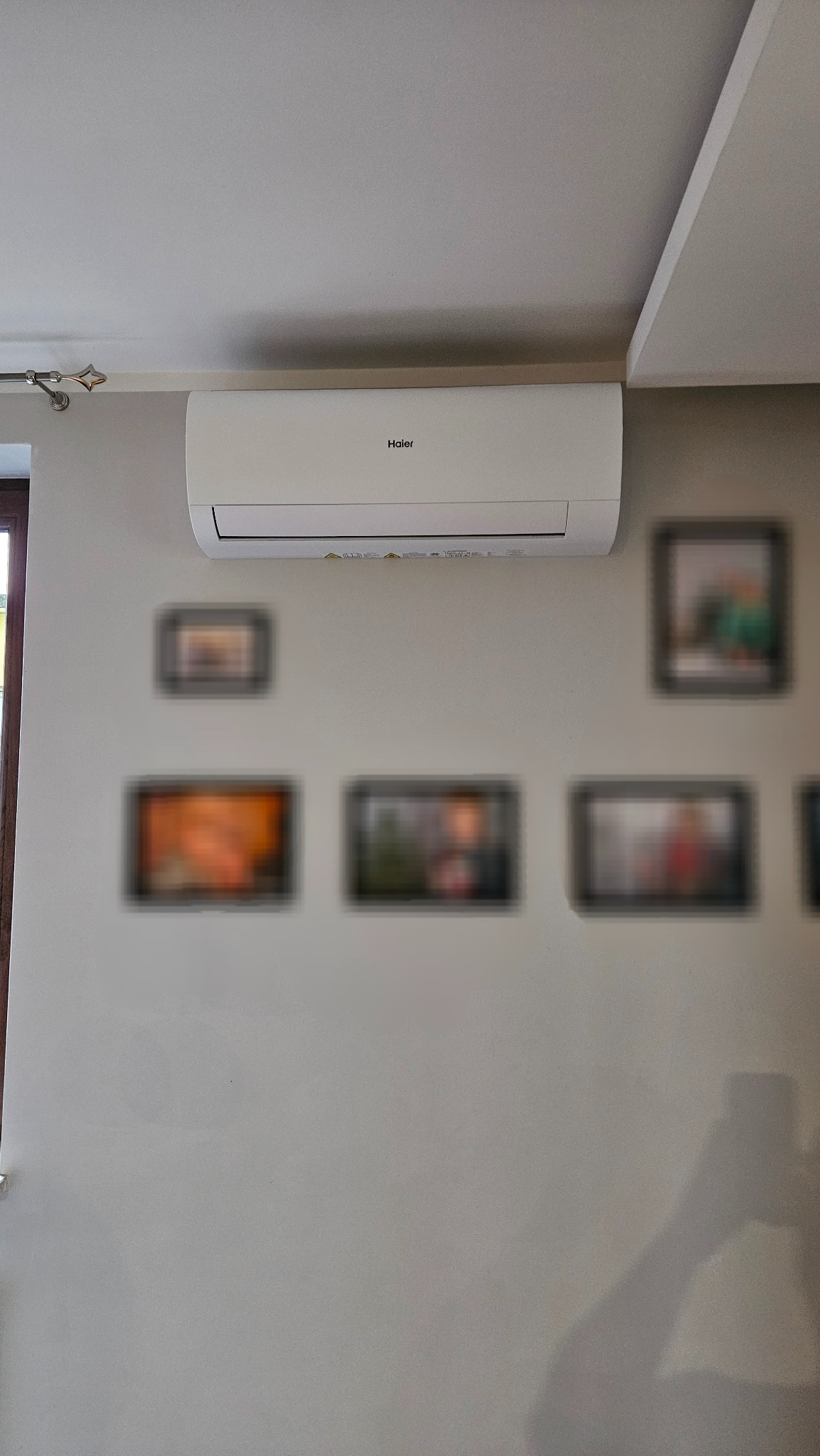 Biały klimatyzator Haier zamontowany na ścianie w pokoju, pod sufitem, z dekoracyjnymi obrazkami w ramkach poniżej. Widoczny fragment okna i cień osoby.