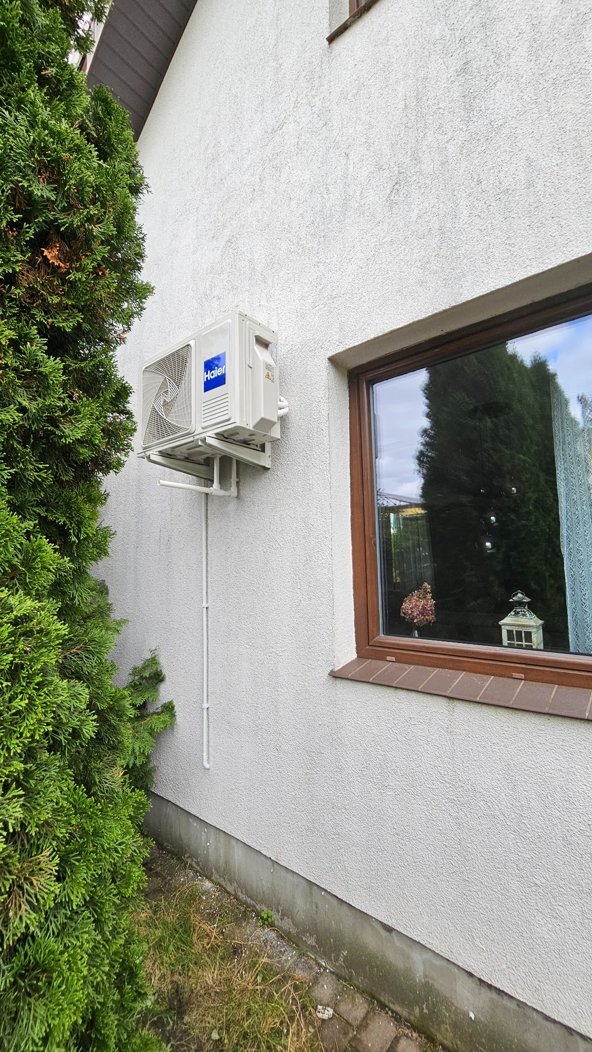 Biała jednostka zewnętrzna klimatyzacji Haier zamontowana na elewacji domu obok okna, z widoczną instalacją i zielonym krzewem z boku.