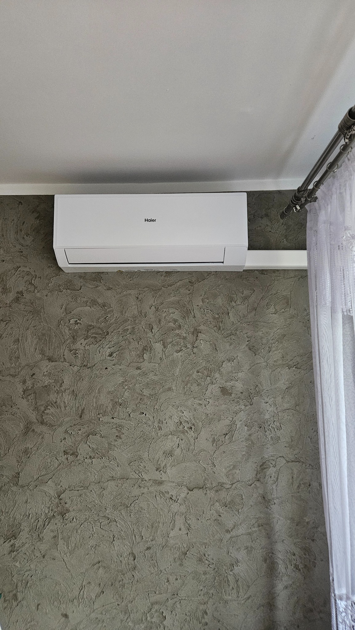 Biały klimatyzator Haier zamontowany na szarej, teksturowanej ścianie. Widoczny fragment białej zasłony i karnisza. Ujęcie z dołu, widoczny fragment sufitu.