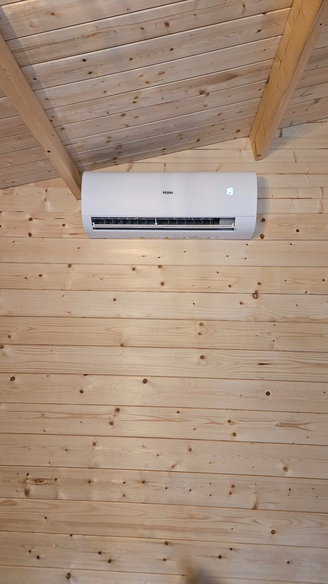 Biały klimatyzator Haier zamontowany na ścianie z jasnych, drewnianych paneli w pomieszczeniu z drewnianym sufitem. Widoczny wyświetlacz temperatury.