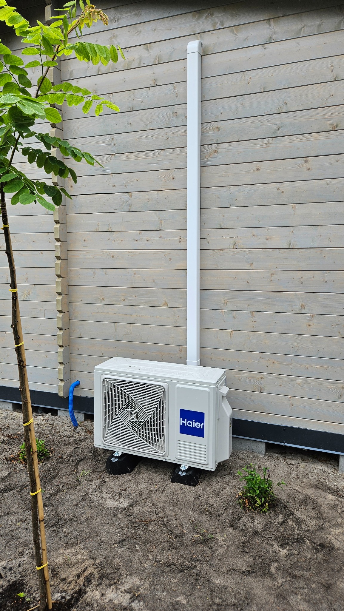 Biały klimatyzator Haier zamontowany na zewnątrz drewnianego budynku z białą rurą odprowadzającą, widoczny fragment drzewa po lewej stronie.