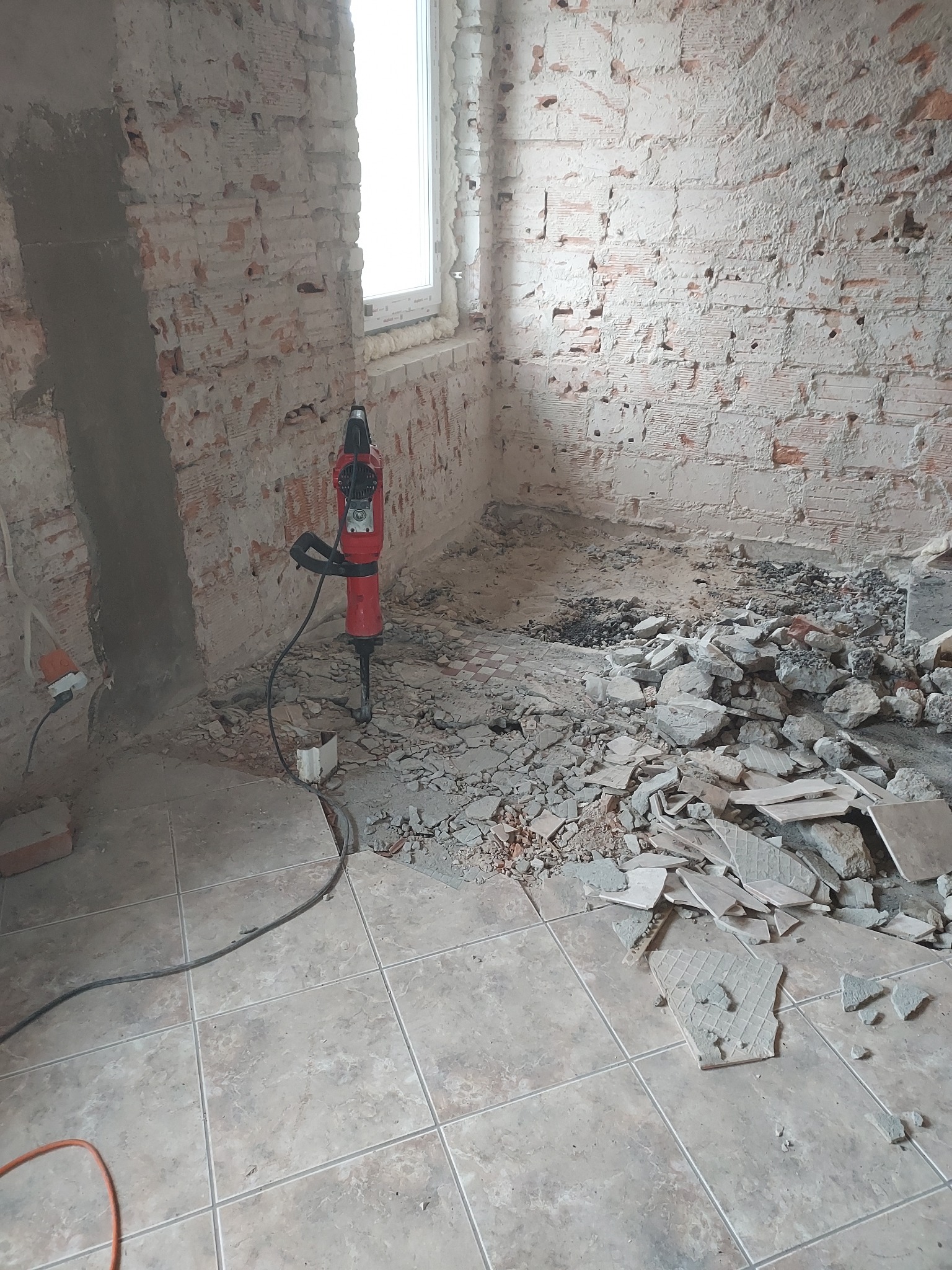 Ceglana ściana po skuciu tynku, gruz na podłodze z płytek, czerwony młot pneumatyczny. Etap prac wyburzeniowych w pomieszczeniu z oknem.