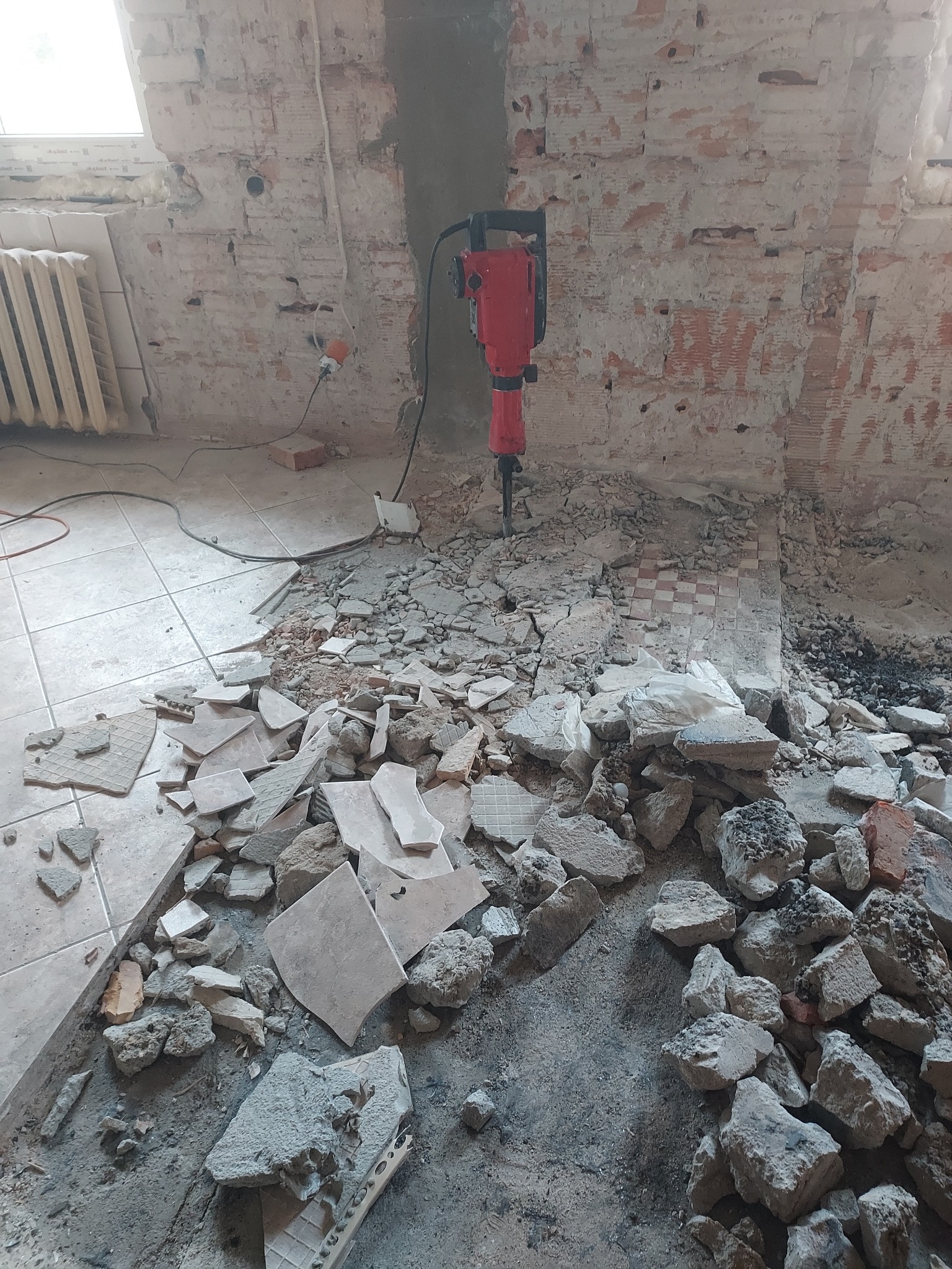 Czerwony młot udarowy rozbija starą podłogę z płytek w pomieszczeniu z odsłoniętą cegłą. Gruz i fragmenty płytek na podłodze. Widoczne okno i grzejnik.