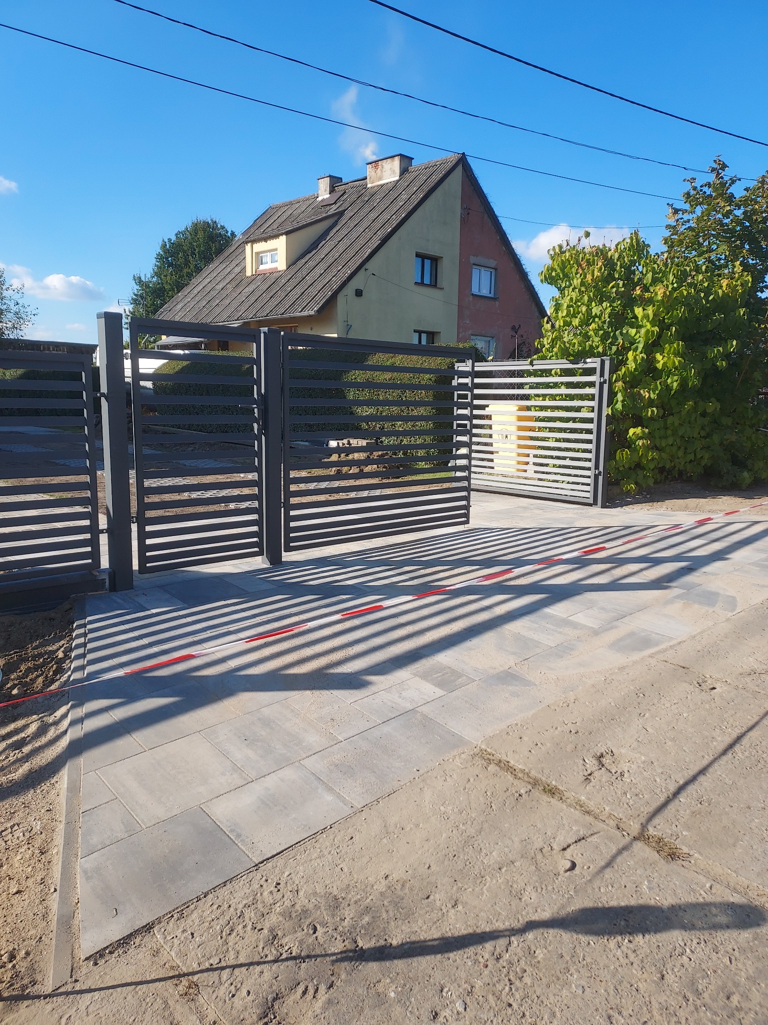 Nowoczesne, poziome ogrodzenie panelowe w kolorze grafitowym, z bramą wjazdową i furtką, na tle domu i kostki brukowej. Widoczne zabezpieczenia budowy.