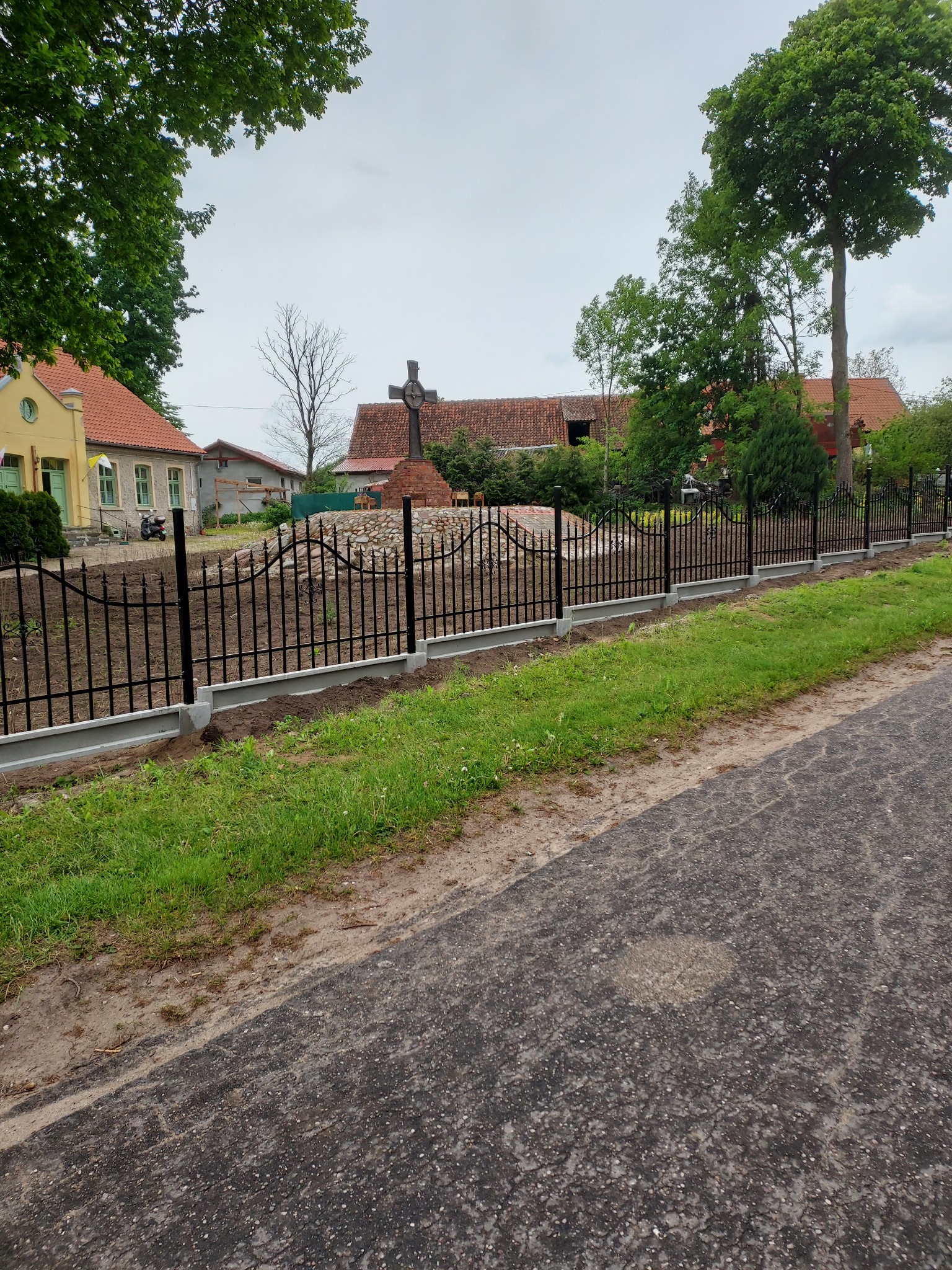 Nowoczesne, czarne ogrodzenie metalowe z betonową podmurówką, otaczające teren z krzyżem. Widok z asfaltowej drogi, w tle budynki i zieleń.