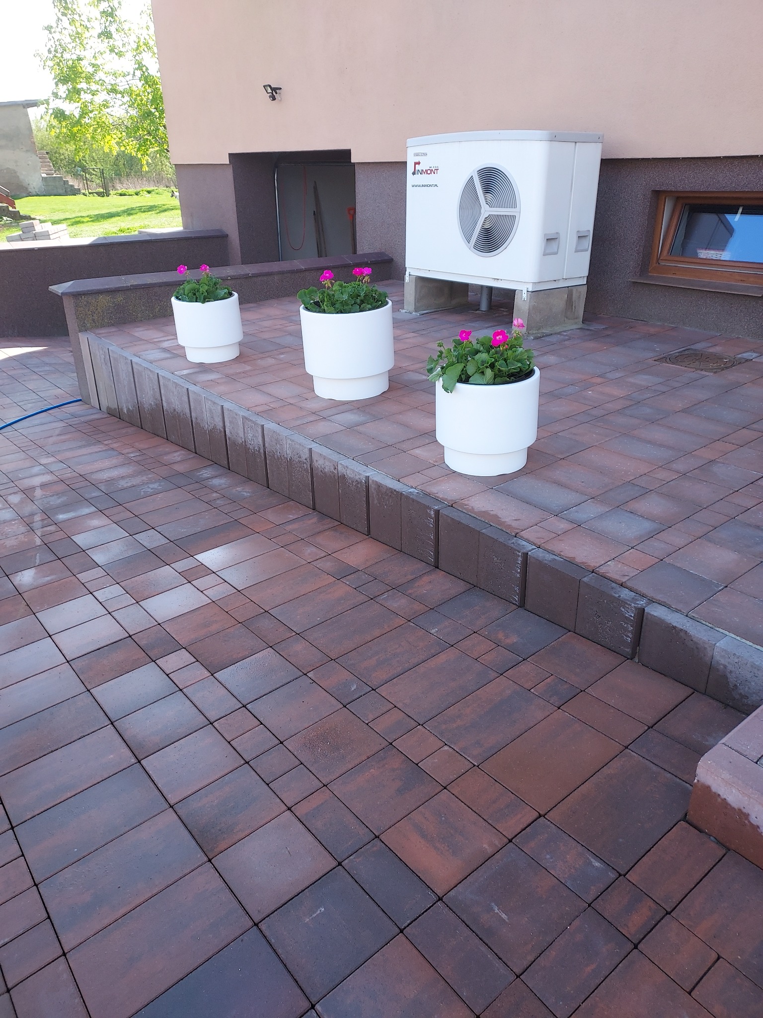 Ułożona kostka brukowa w odcieniach brązu, z trzema białymi donicami z różowymi kwiatami. W tle budynek z pompą ciepła. Widok na taras.