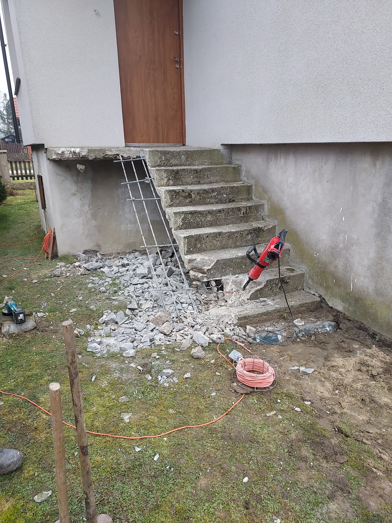Demontaż betonowych schodów wejściowych do domu. Widoczny gruz, zbrojenie i młot pneumatyczny. Prace rozbiórkowe w toku, teren wokół częściowo uprzątnięty.
