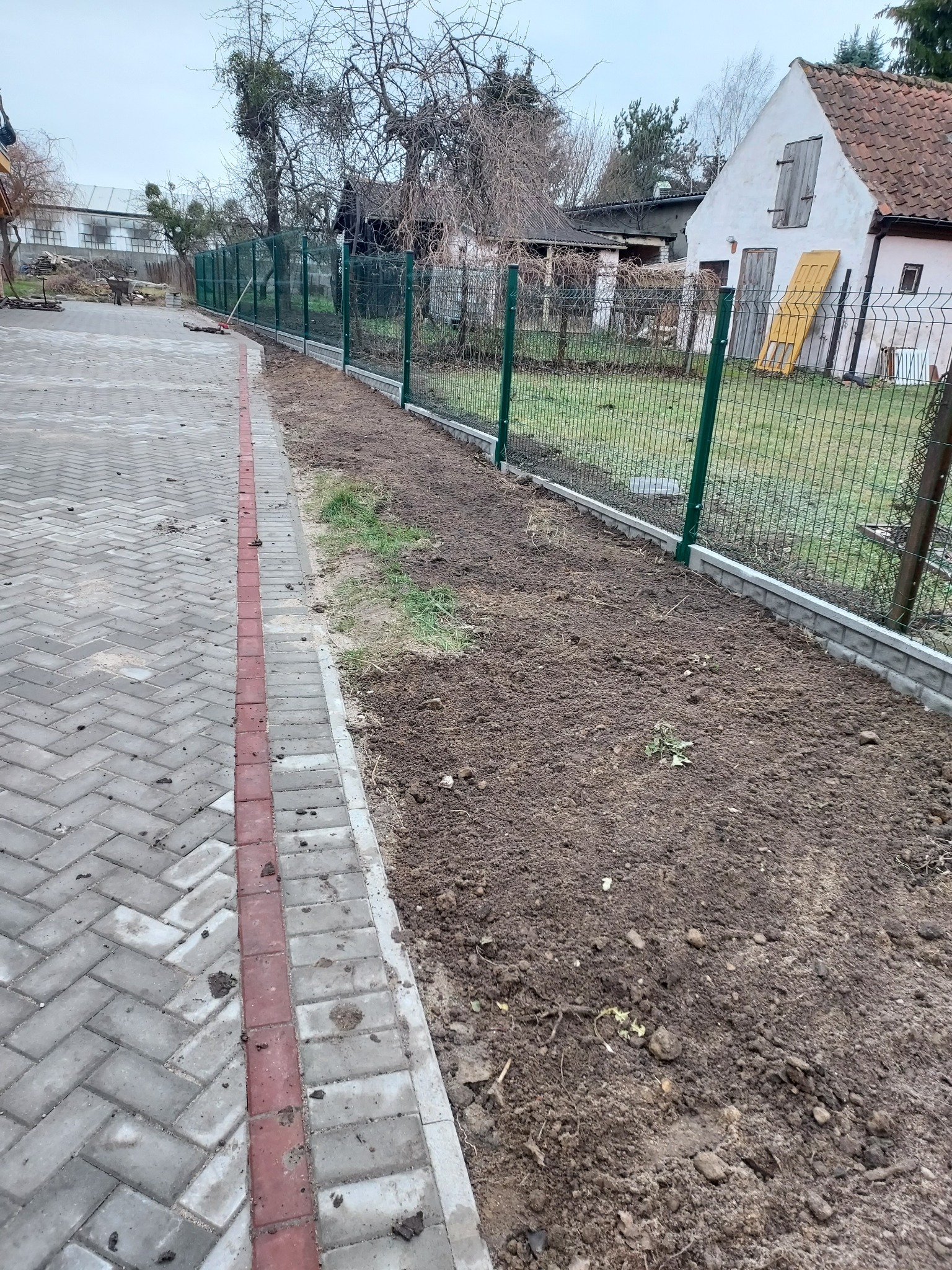 Nowo postawione ogrodzenie z siatki w zielonej ramie, oddzielające posesję od chodnika z kostki. Przygotowany pas ziemi pod nasadzenia wzdłuż ogrodzenia.