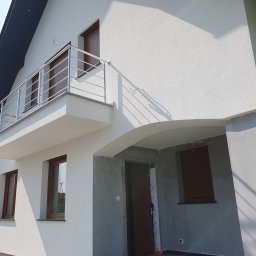 DARPROTECT DARIUSZ MARSZAŁEK - Biała elewacja domu z balkonem i brązowymi oknami. Część elewacji w szarym tynku. Widoczne schody wejściowe i fragment dachu. Ujęcie w słoneczny dzień.