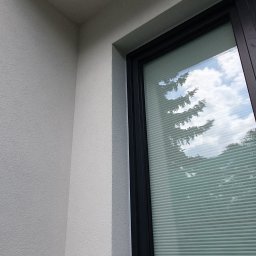 DARPROTECT DARIUSZ MARSZAŁEK - Fragment elewacji budynku z oknem z żaluzjami, odbijającymi drzewa i niebo. Ściana o jasnej, ziarnistej fakturze. Czarna rama okienna.