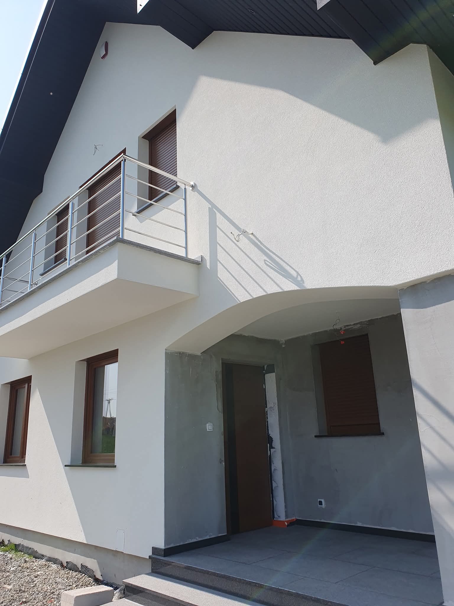 Biała elewacja domu z balkonem i brązowymi oknami. Część elewacji w szarym tynku. Widoczne schody wejściowe i fragment dachu. Ujęcie w słoneczny dzień.