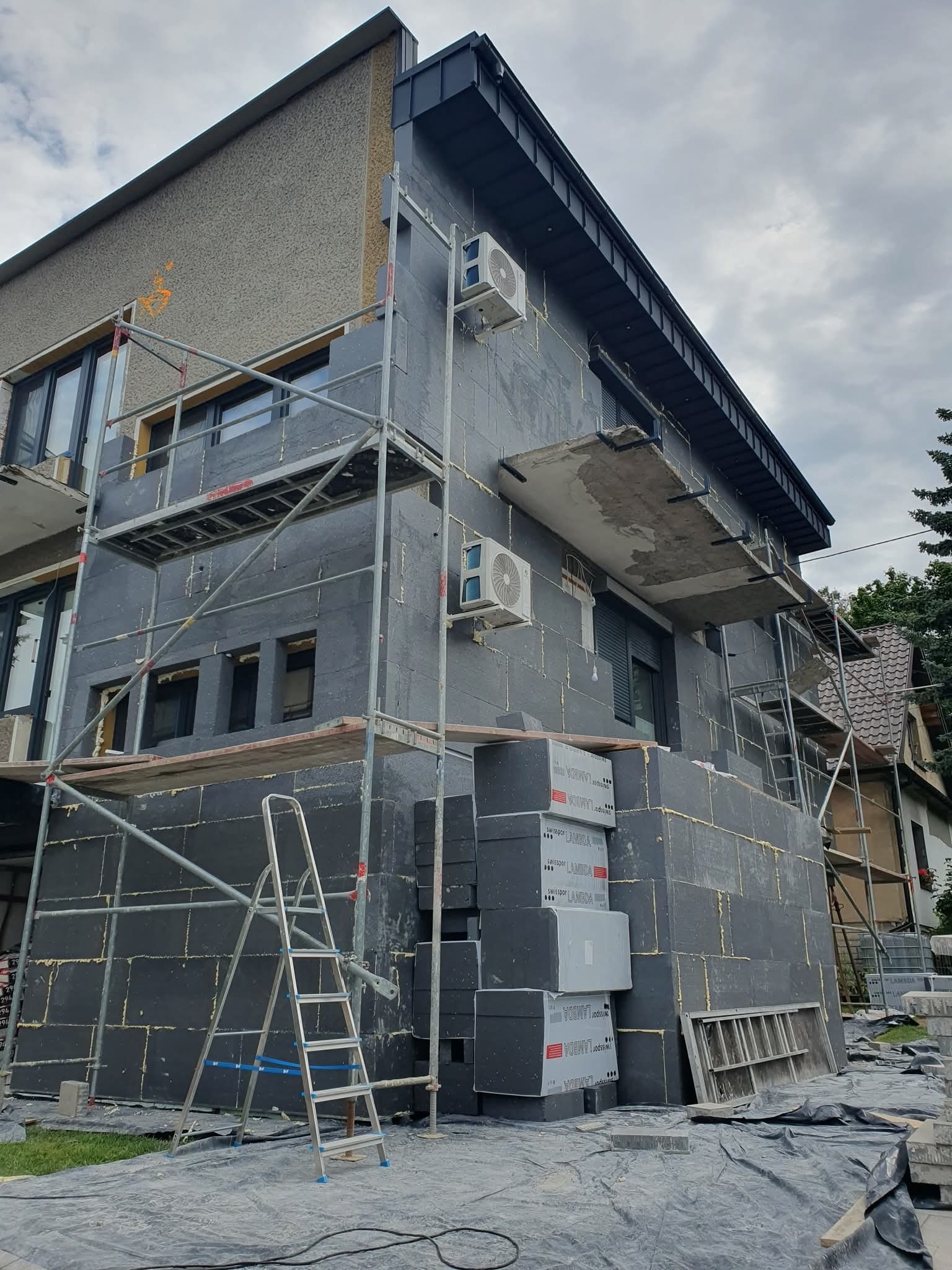 Ocieplanie elewacji budynku mieszkalnego styropianem grafitowym, widoczne rusztowania i drabina. Częściowo ocieplona ściana z zamontowanymi klimatyzatorami.