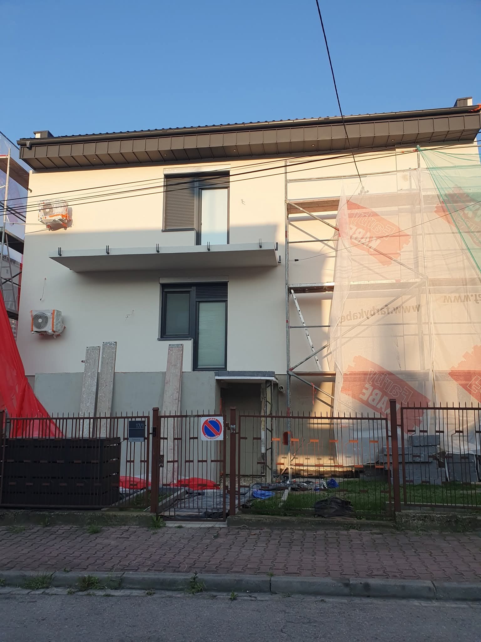 Dom jednorodzinny w trakcie ocieplania elewacji, widoczne rusztowania z siatką ochronną, częściowo otynkowana ściana, elementy klimatyzacji i ogrodzenie.