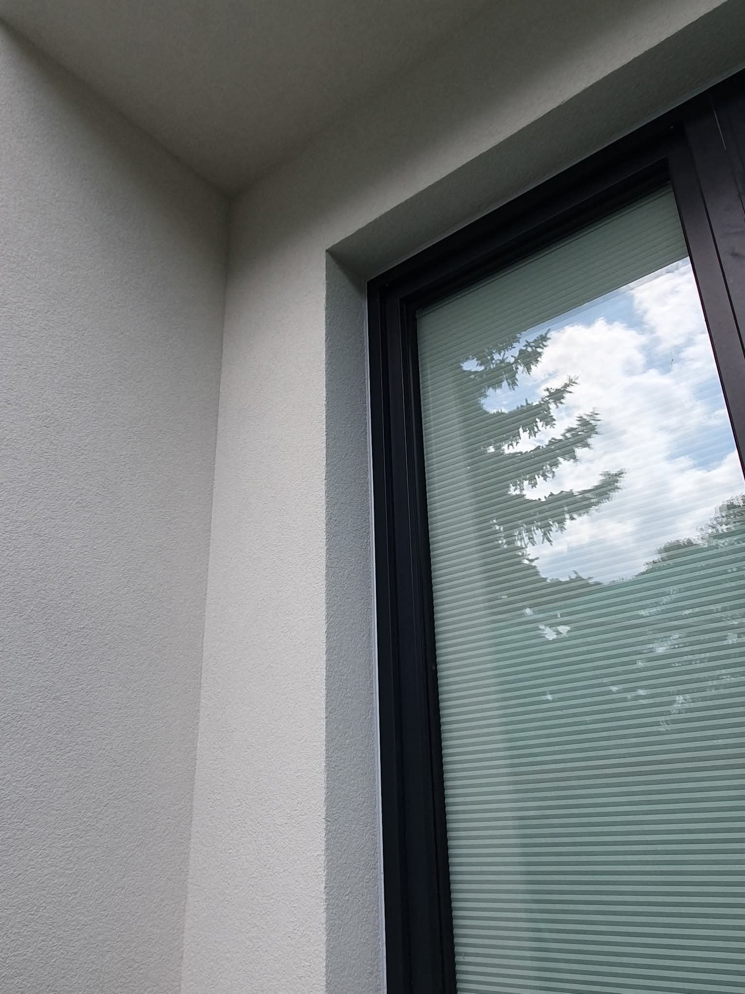 Fragment elewacji budynku z oknem z żaluzjami, odbijającymi drzewa i niebo. Ściana o jasnej, ziarnistej fakturze. Czarna rama okienna.