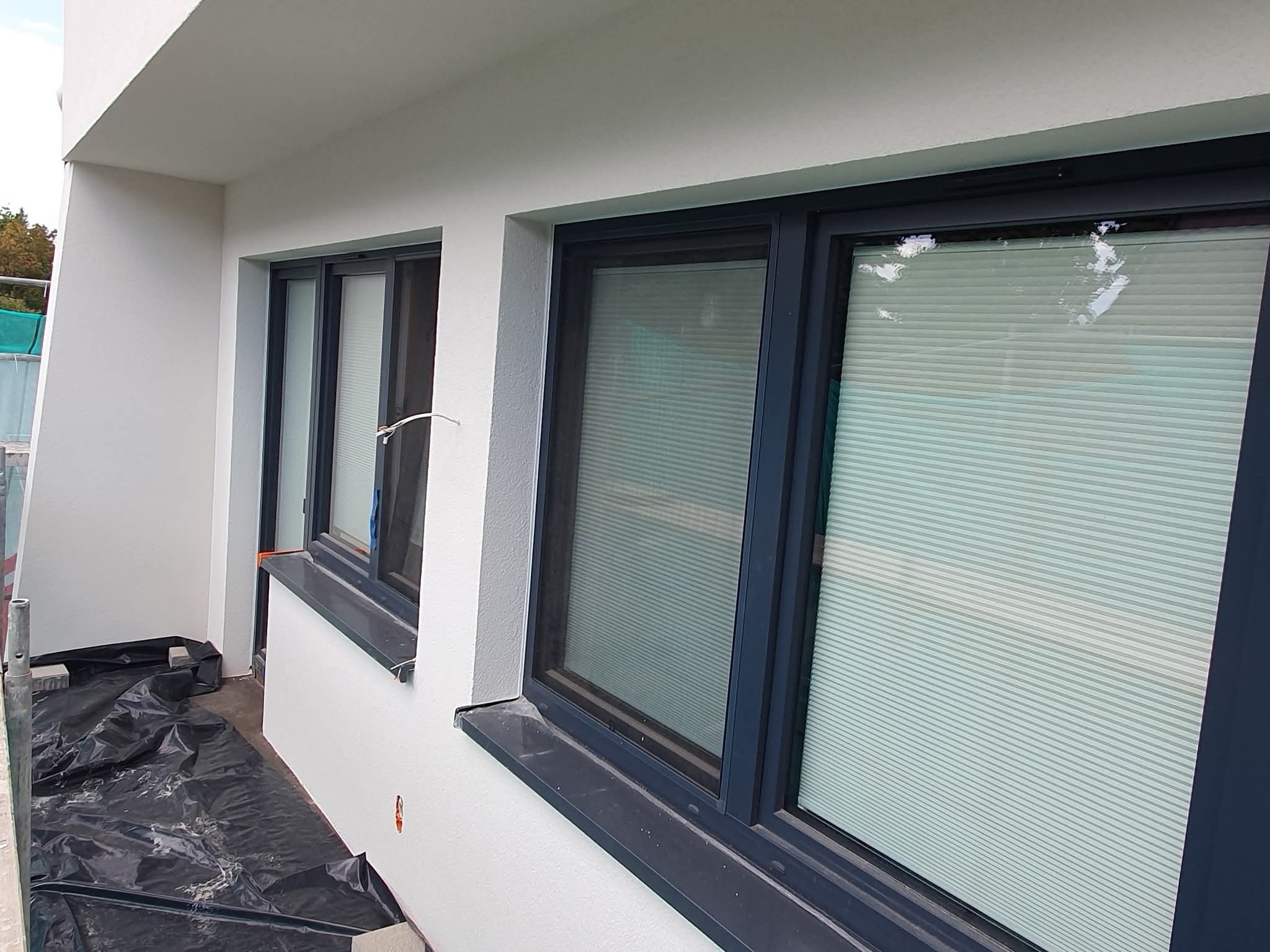 Biała elewacja budynku po malowaniu, widoczne okna z szarymi ramami i zasłonięte żaluzjami. Na podłożu folia ochronna, ślady prac malarskich.