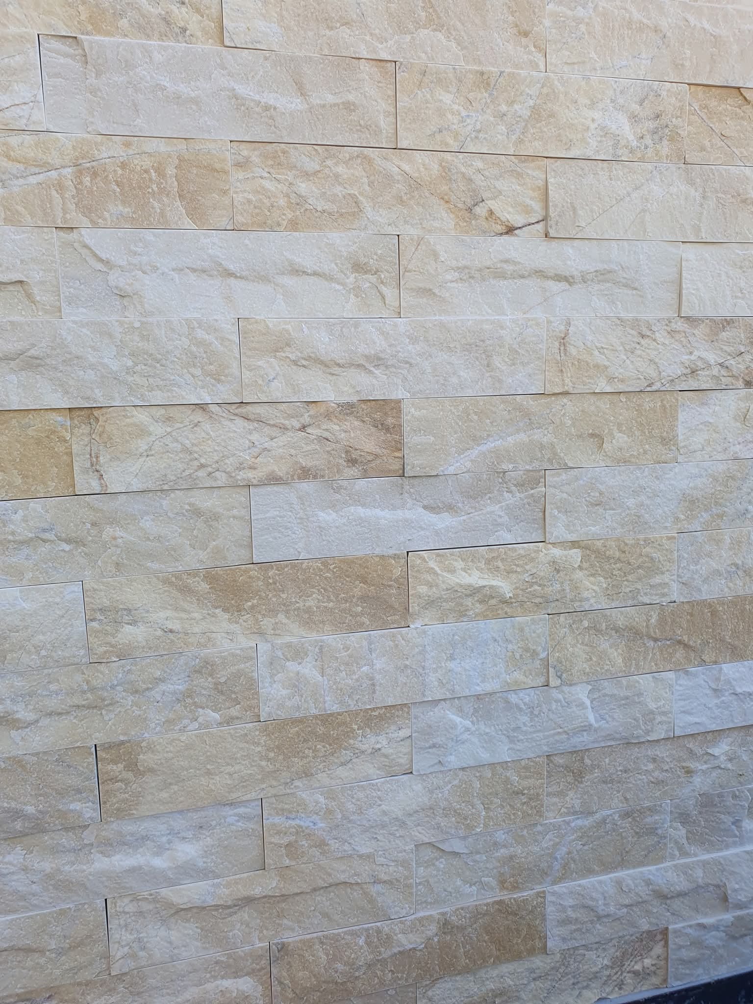 Ściana wykończona beżowymi, prostokątnymi płytkami imitującymi kamień naturalny, ułożonymi w regularny wzór z przesunięciem. Widoczna faktura i delikatne cieniowanie płytek.