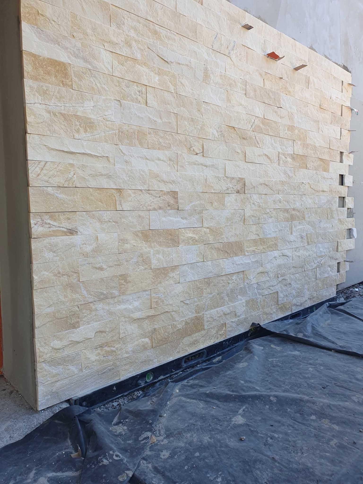 Ściana wykończona beżową okładziną kamienną, widoczne detale faktury i fugi. Częściowo ułożona, z widocznymi elementami montażowymi i folią ochronną u dołu.