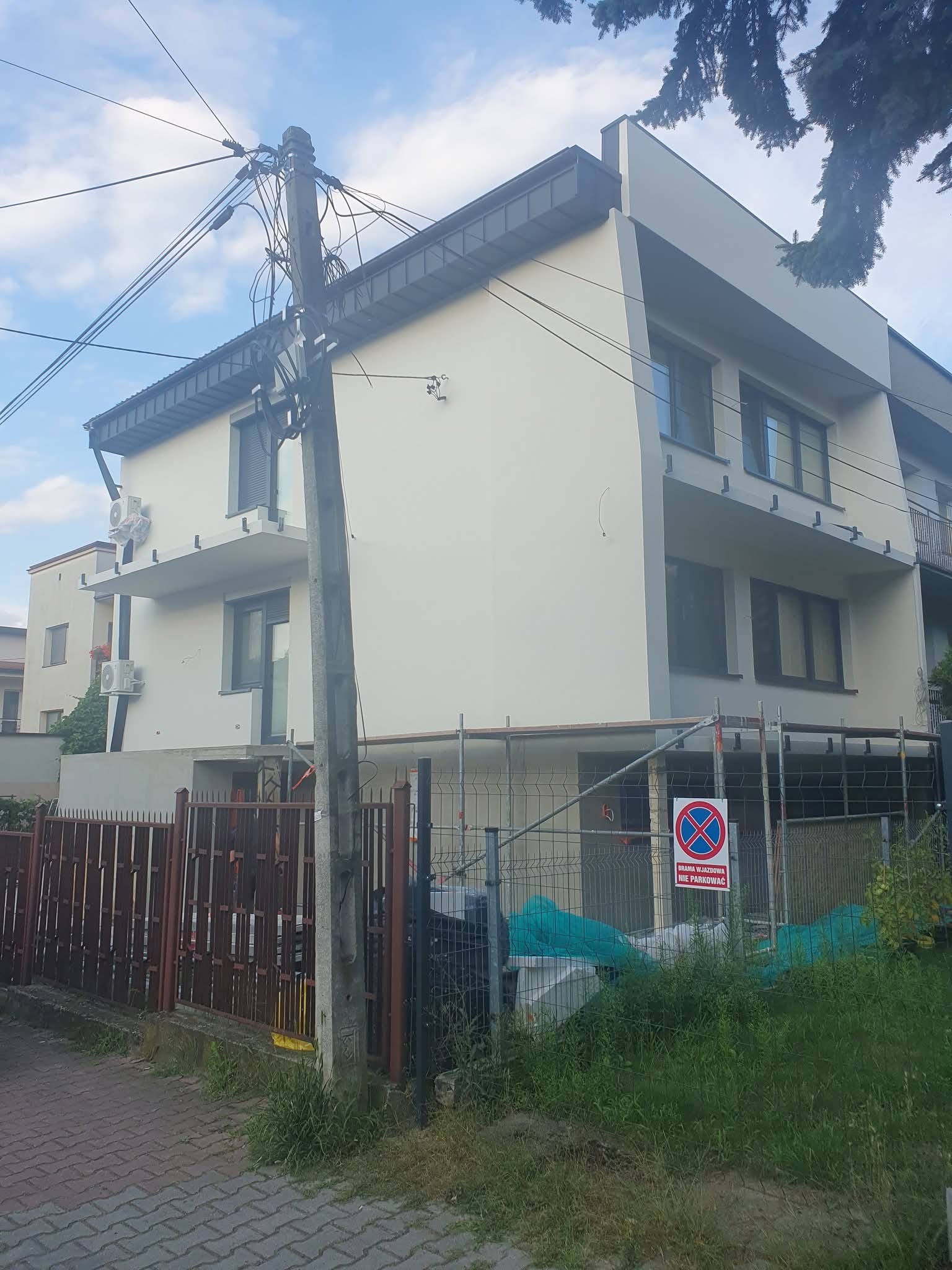 Dwupiętrowy budynek po remoncie elewacji, z widocznymi rusztowaniami i znakiem zakazu parkowania. Świeżo pomalowana fasada w odcieniach bieli i szarości.