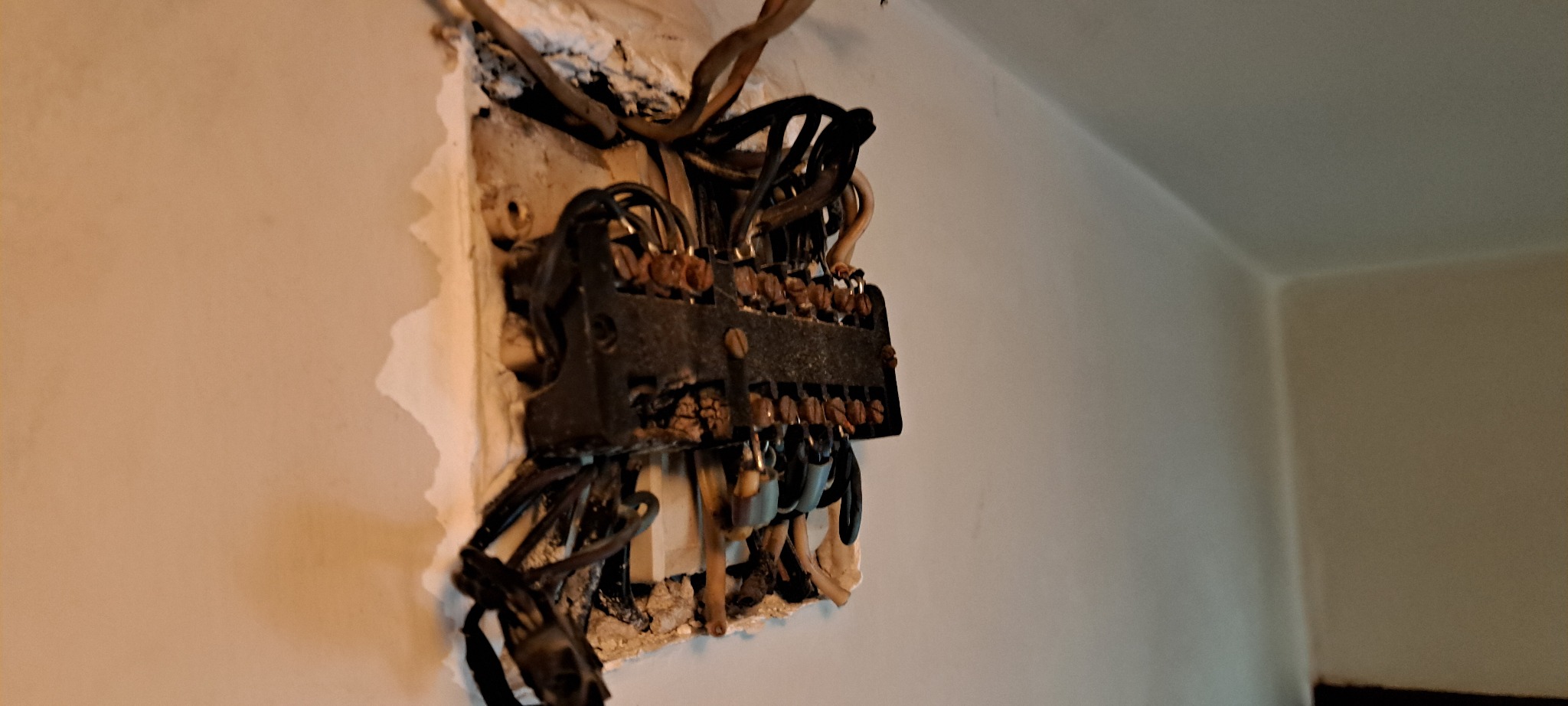 Uszkodzona, stara instalacja elektryczna z wypalonymi przewodami, widoczna na tle ściany. Zniszczona skrzynka bezpiecznikowa z licznymi kablami.