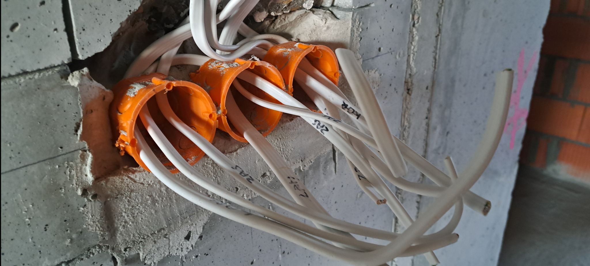 Fragment ściany z trzema pomarańczowymi puszkami instalacyjnymi, z których wychodzą białe kable elektryczne z naniesionymi oznaczeniami. Surowy beton i cegła w tle.