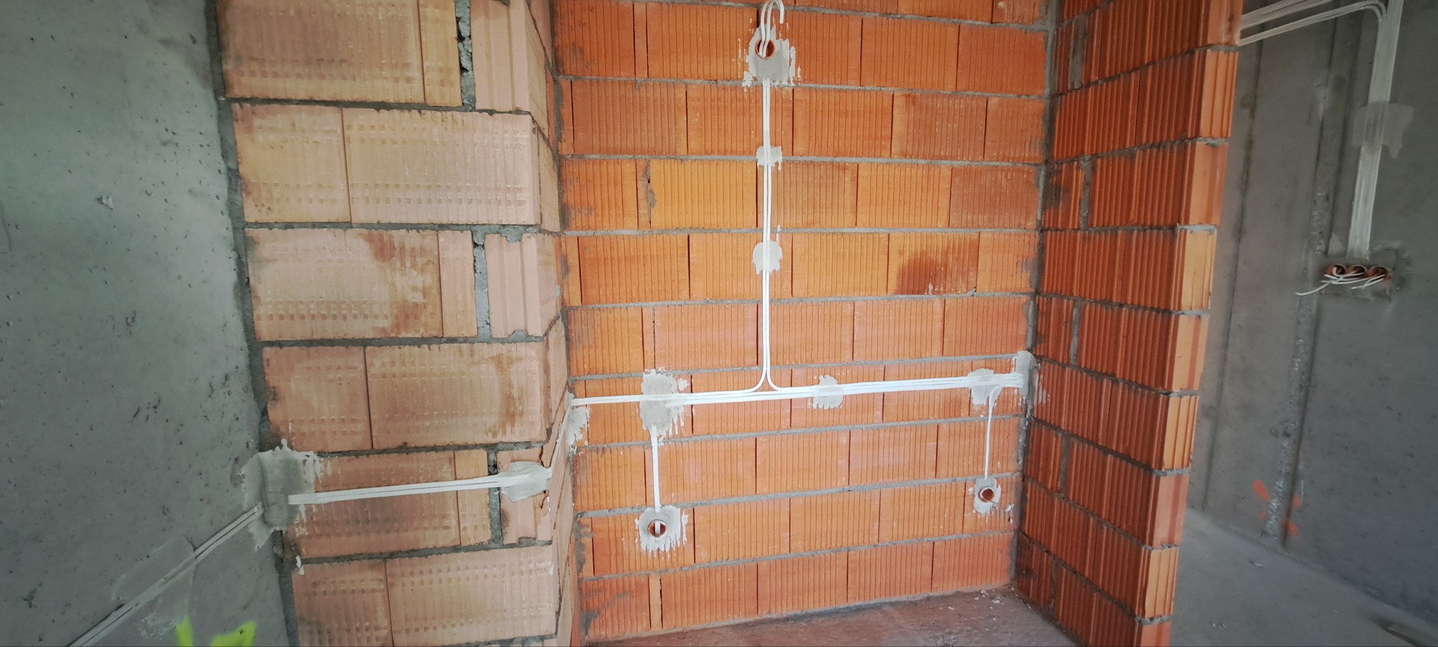 Wewnętrzna instalacja elektryczna w surowym stanie: białe przewody przymocowane do ściany z czerwonej cegły dziurawki, widoczne puszki pod gniazdka i kable na ścianie betonowej.
