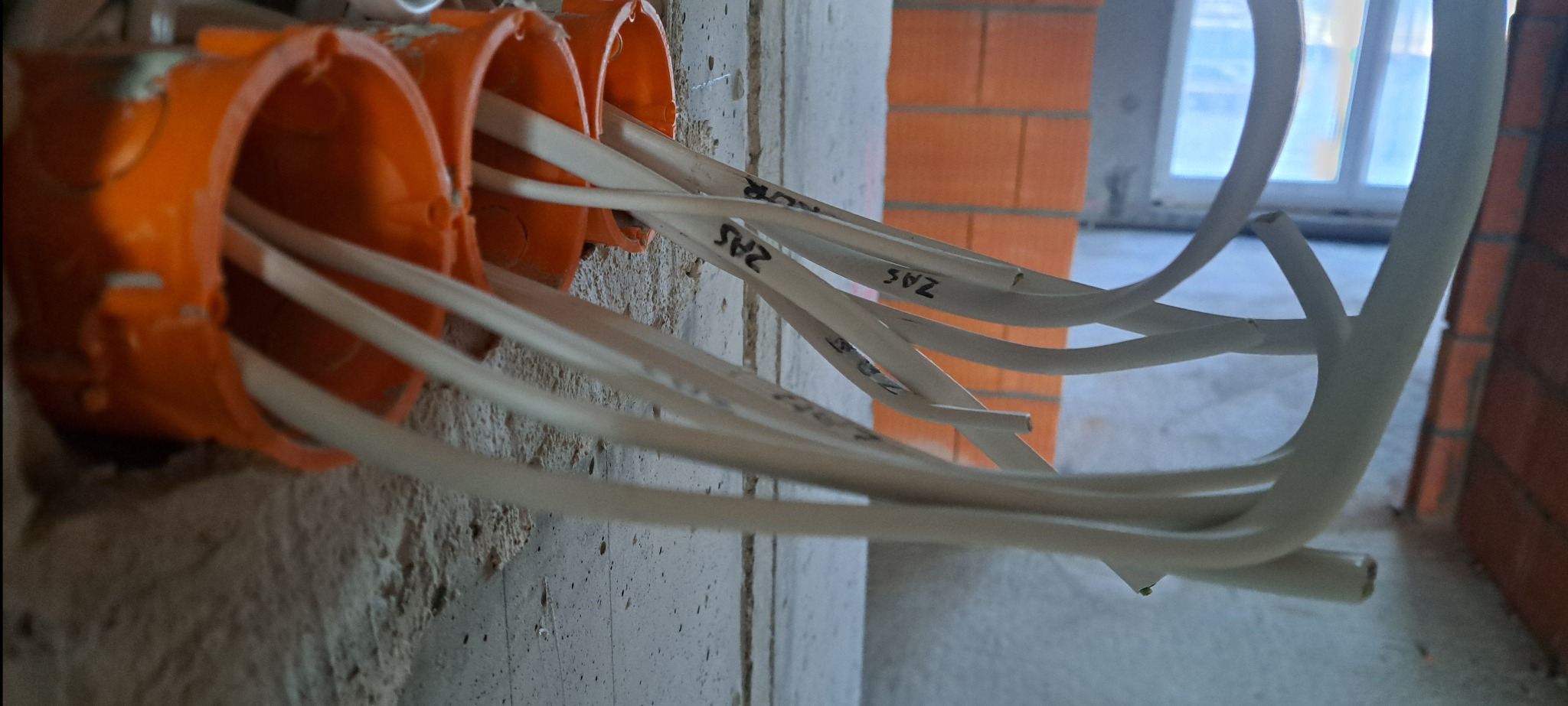 Pomarańczowe puszki instalacyjne z białymi przewodami oznaczonymi napisami, zamontowane w surowej ścianie z betonu i cegieł. Widok z bliska na instalację elektryczną.